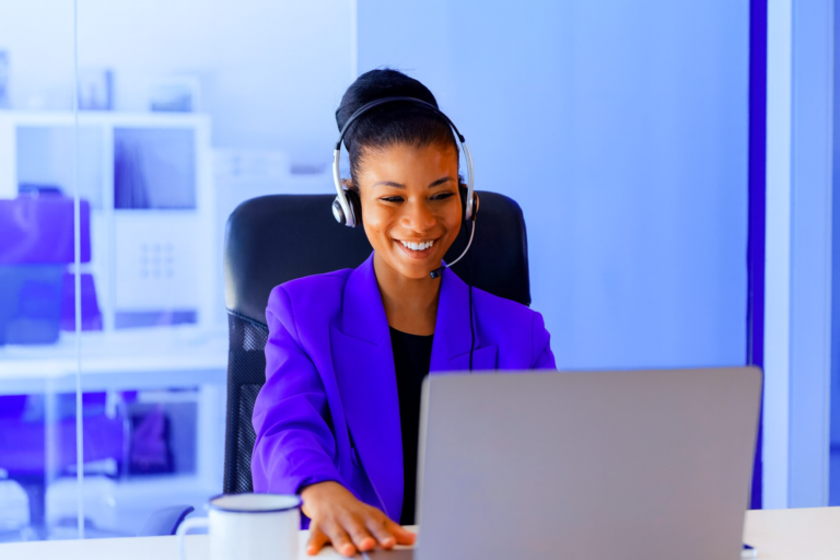 Mulher sorridente com headset atendendo cliente no computador.