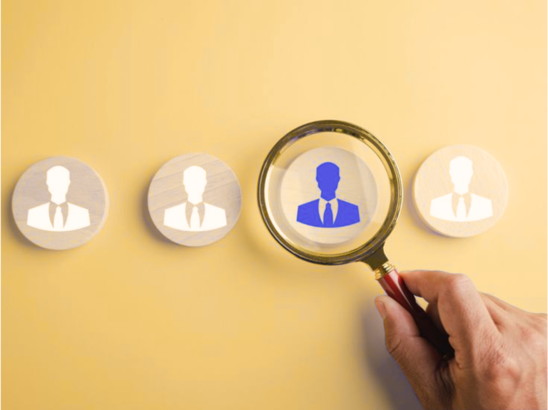 Mão segurando lupa sobre um ícone azul de pessoa entre outros ícones brancos, em fundo amarelo.