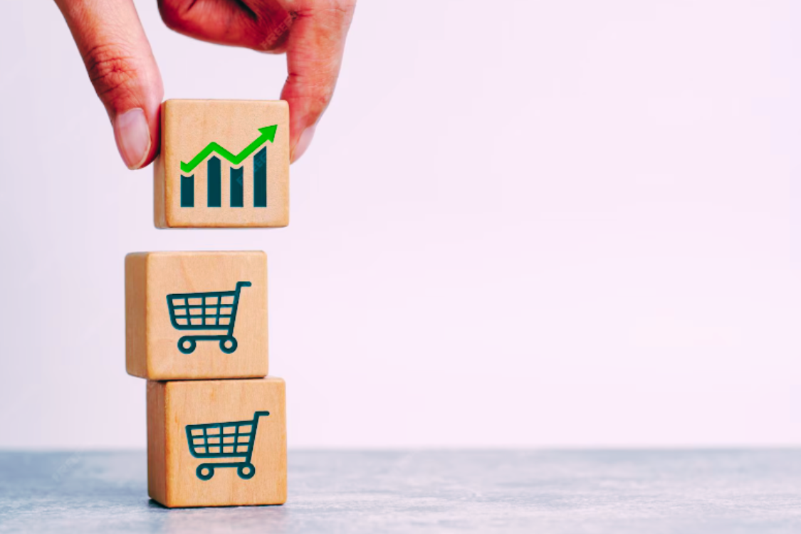 Mão colocando um bloco de madeira com ícone de gráfico crescente acima de dois blocos com ícones de carrinho de compras.