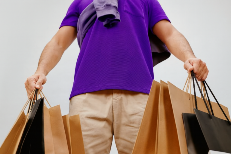 Texto alternativo (alt text): Pessoa com várias sacolas de compras nas mãos, vestindo camisa roxa e calça bege.