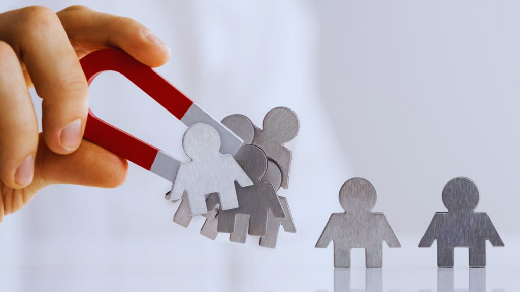 Mão segurando um ímã em formato de ferradura atraindo figuras metálicas que representam pessoas, simbolizando atração e captação de clientes.